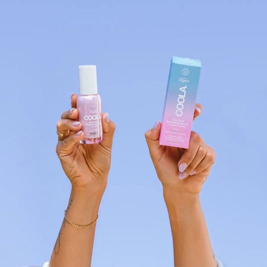COOLA Dew Good Illuminating Serum SPF 30 holdt i hænder mod lyseblå baggrund